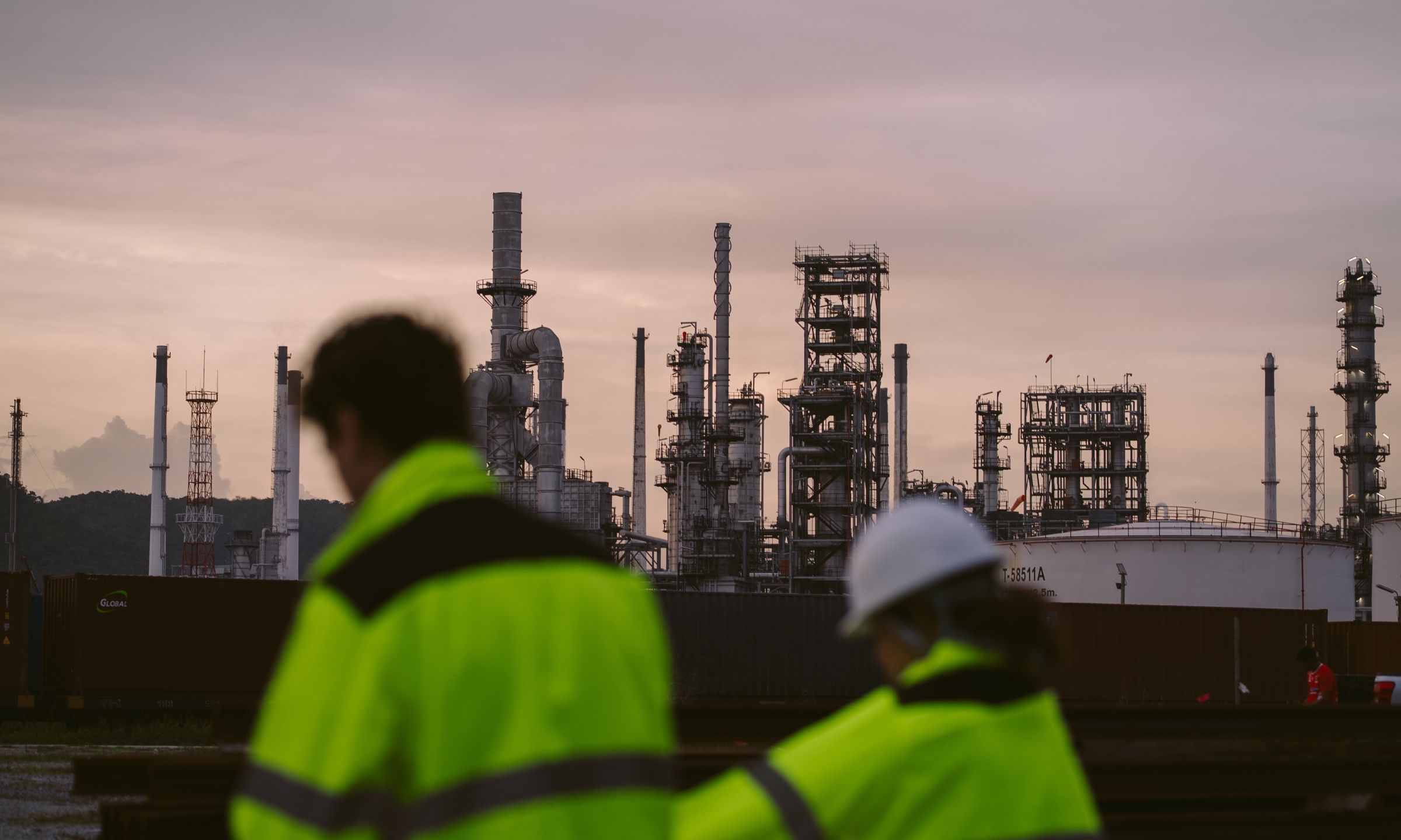 Workers in high-visibility jackets walking past an industrial refinery with large processing towers and pipelines at dusk.
