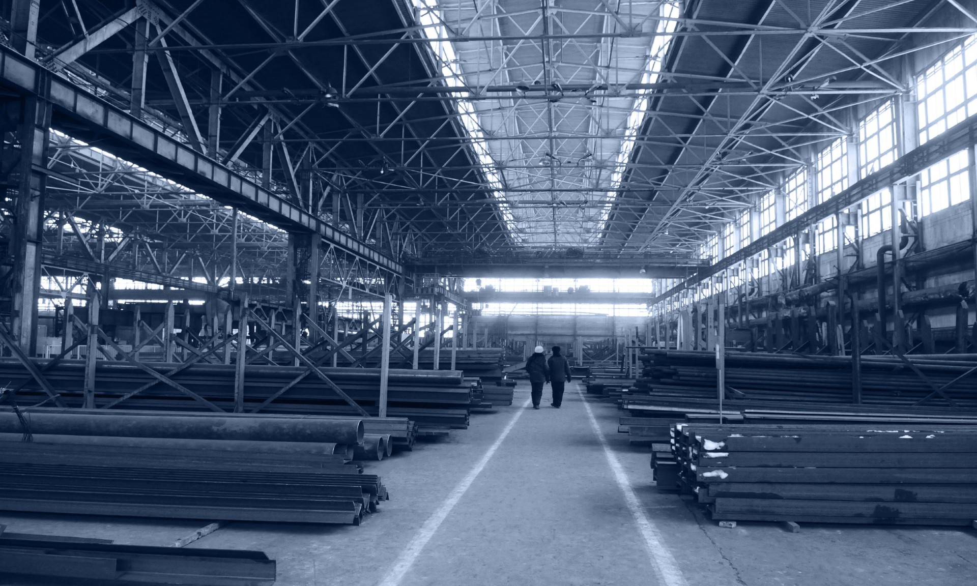 Industrial warehouse interior with rows of stacked steel beams and pipes, overhead metal framework, and two workers walking down a central aisle.