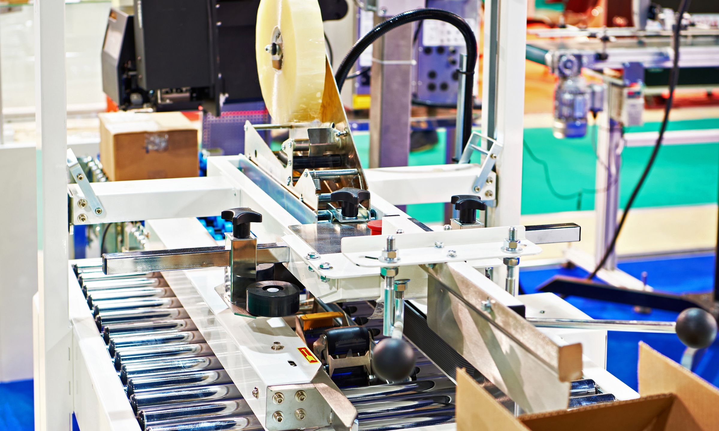 Automated packaging line applying tape to cardboard boxes on a conveyor system in a manufacturing facility.